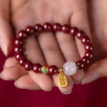 A string of deep red cinnabar beads featuring a luminous white stone and a dangling gold wealth amulet with Chinese characters.