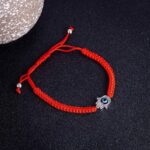 A vibrant red braided cord bracelet featuring an adjustable sliding knot closure and a silver-toned Hamsa hand charm with a central blue Evil Eye.