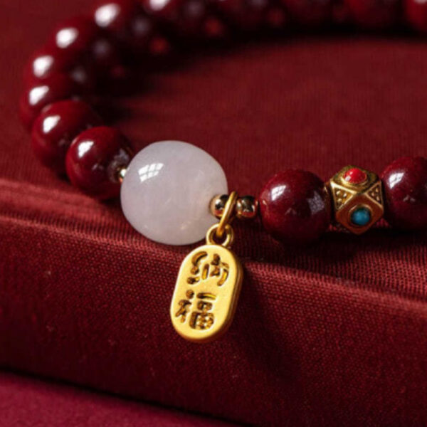 A string of deep red cinnabar beads featuring a luminous white stone and a dangling gold wealth amulet with Chinese characters.