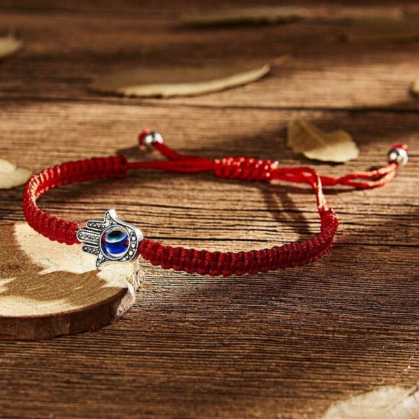 A vibrant red braided cord bracelet featuring an adjustable sliding knot closure and a silver-toned Hamsa hand charm with a central blue Evil Eye.