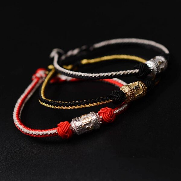 A braided red and gold cord bracelet featuring an engraved golden mantra bead secured by traditional sacred knots against a dark wooden background.