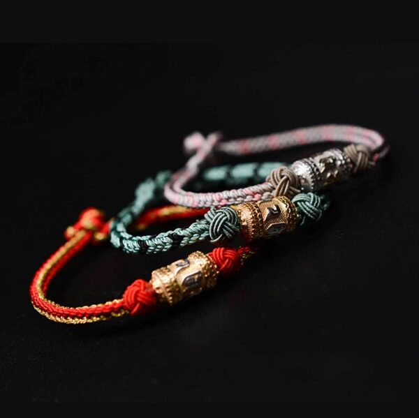 A braided red and gold cord bracelet featuring an engraved golden mantra bead secured by traditional sacred knots against a dark wooden background.