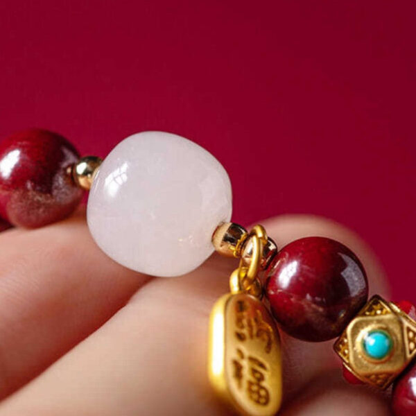 A string of deep red cinnabar beads featuring a luminous white stone and a dangling gold wealth amulet with Chinese characters.