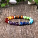 A colorful beaded bracelet showcasing seven distinct gemstone hues separated by textured silver spacers on a rustic wood background.