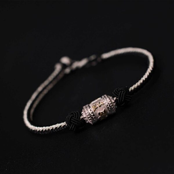 A braided red and gold cord bracelet featuring an engraved golden mantra bead secured by traditional sacred knots against a dark wooden background.