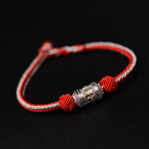 A braided red and gold cord bracelet featuring an engraved golden mantra bead secured by traditional sacred knots against a dark wooden background.
