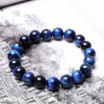 Polished round beads of deep blue Tiger's Eye with striking chatoyancy rest on a textured monochrome surface.