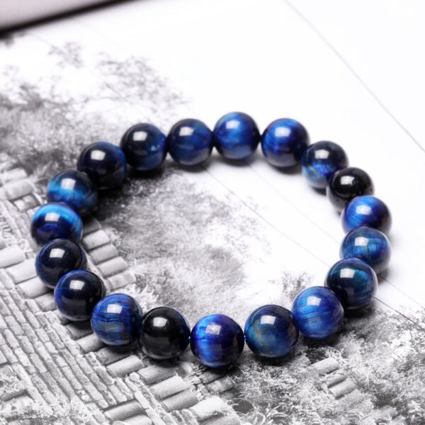 Polished round beads of deep blue Tiger's Eye with striking chatoyancy rest on a textured monochrome surface.