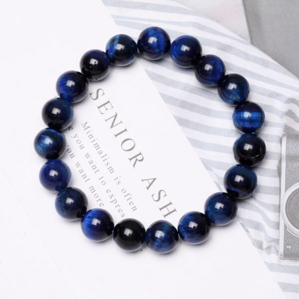 Polished round beads of deep blue Tiger's Eye with striking chatoyancy rest on a textured monochrome surface.
