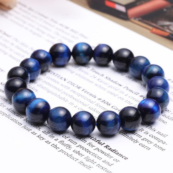 Polished round beads of deep blue Tiger's Eye with striking chatoyancy rest on a textured monochrome surface.