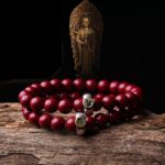 Glossy deep red cinnabar beads strung together with metallic guardian deity charms, displayed on rustic wood before a golden Buddha statue.