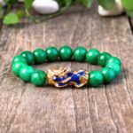 Vibrant green jade beads encircle a striking gold and blue enamel Pixiu charm set against a rustic wooden background.