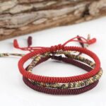 Three intricately braided rope bracelets in vibrant red, deep maroon, and multi-colored threads featuring adjustable sliding knots against a natural wood background.