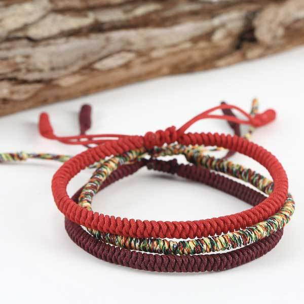 Three intricately braided rope bracelets in vibrant red, deep maroon, and multi-colored threads featuring adjustable sliding knots against a natural wood background.