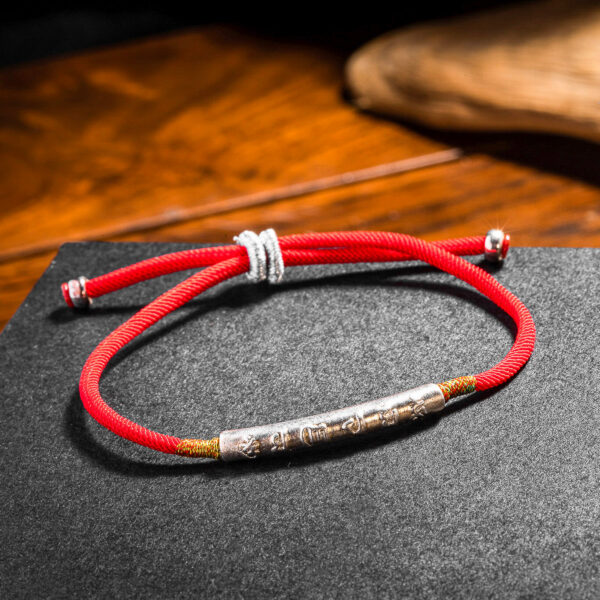 A vibrant red braided cord bracelet featuring an engraved gold-tone cylindrical bead and adjustable sliding knots.