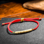 A vibrant red braided cord bracelet featuring an engraved gold-tone cylindrical bead and adjustable sliding knots.