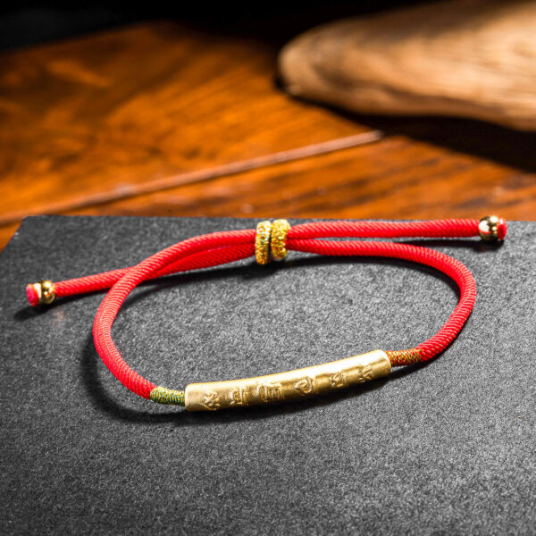 A vibrant red braided cord bracelet featuring an engraved gold-tone cylindrical bead and adjustable sliding knots.