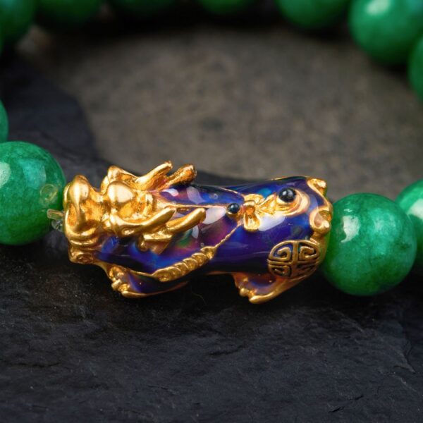 Vibrant green jade beads encircle a striking gold and blue enamel Pixiu charm set against a rustic wooden background.