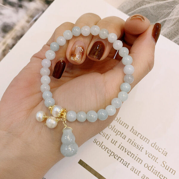 Featuring smooth light green jade beads, this bracelet is elegantly detailed with white pearls and a traditional Wu Lou gourd charm on gold accents.