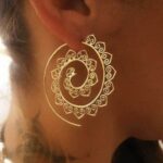 A large gold-toned wire hoop earring featuring an intricate inward spiraling pattern adorned with repeating small heart shapes.