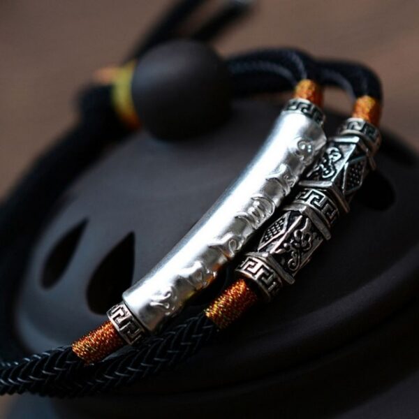 A double-strand black braided cord bracelet featuring a prominent silver cylindrical mantra bar and intricate geometric metal beads with orange gold accents.