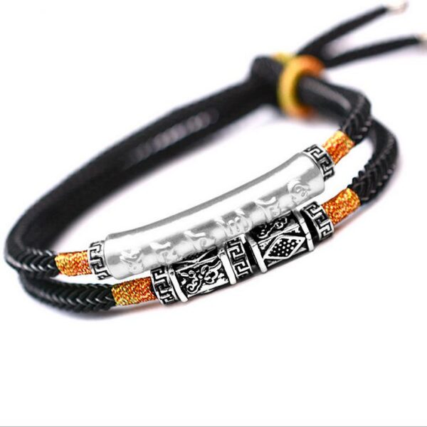 A double-strand black braided cord bracelet featuring a prominent silver cylindrical mantra bar and intricate geometric metal beads with orange gold accents.