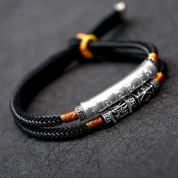 A double-strand black braided cord bracelet featuring a prominent silver cylindrical mantra bar and intricate geometric metal beads with orange gold accents.