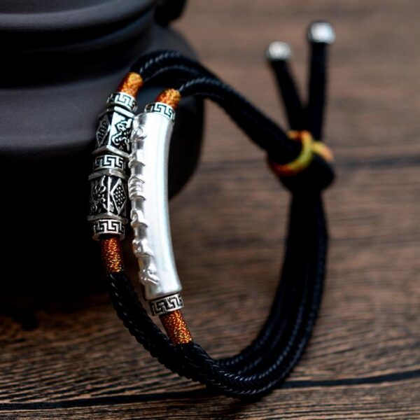 A double-strand black braided cord bracelet featuring a prominent silver cylindrical mantra bar and intricate geometric metal beads with orange gold accents.